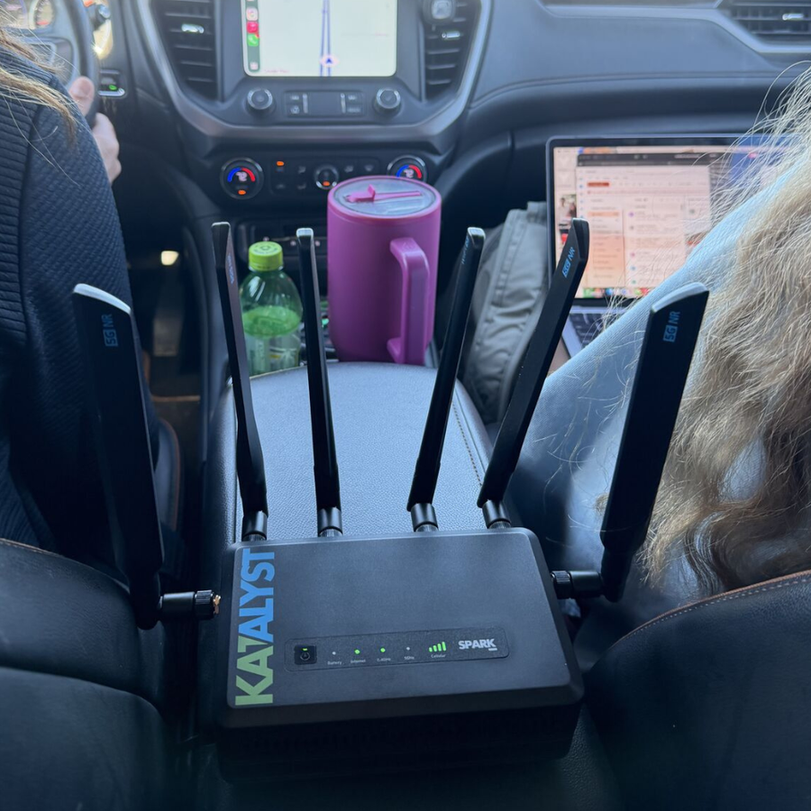 Car interior with a Katalyst Spark router on the dashboard, laptop, and pink mug.