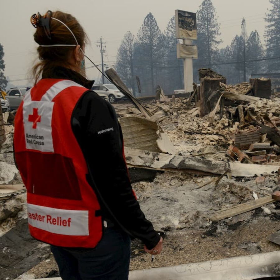 Person wearing an American Red Cross vest in front of a disaster-stricken area with debris and smoke.